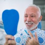 Elderly man sitting in dental care smiling and looking at his dental implants in a hand mirror stock photo