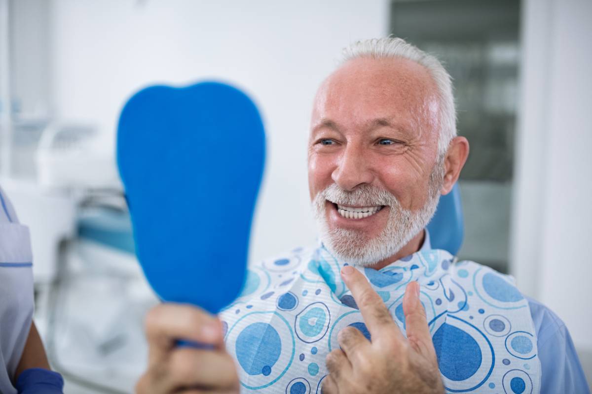 Elderly man sitting in dental care smiling and looking at his dental implants in a hand mirror stock photo